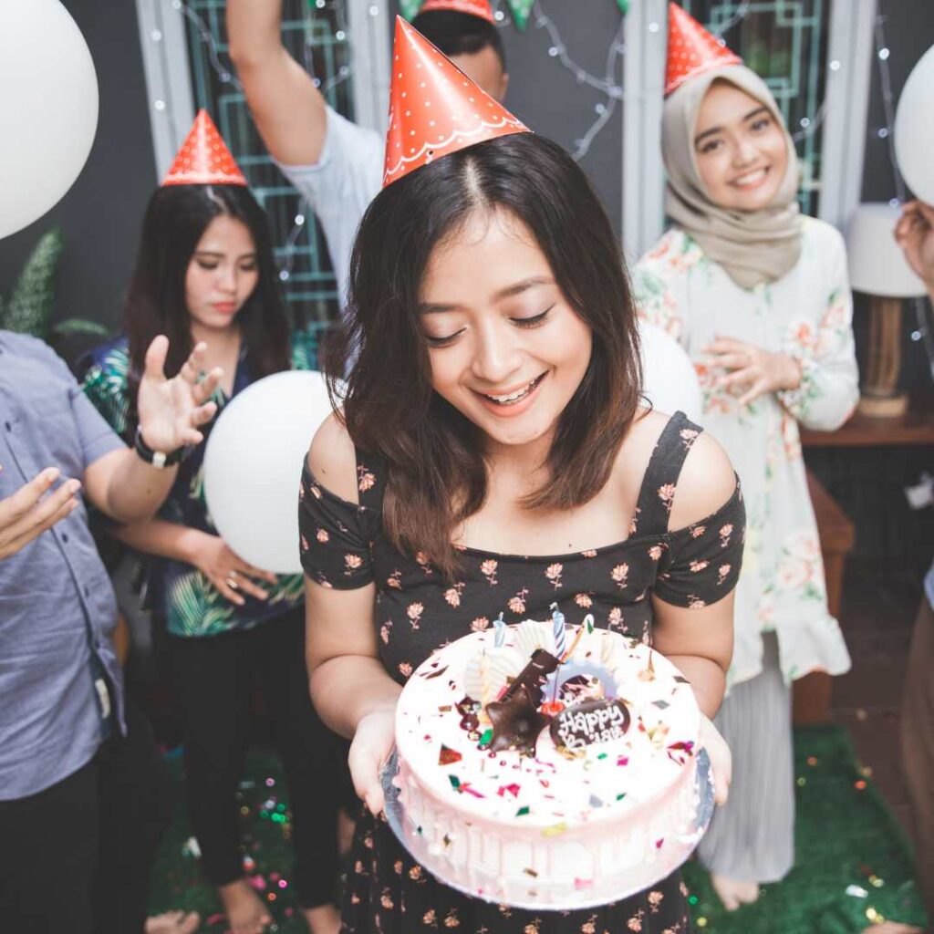 A young woman bringing birthday cake with candles for her friend during a birthday party at home - H